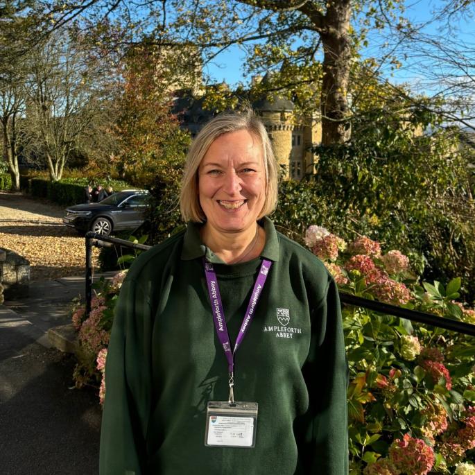 Woman with bobbed blonde hair in green Ampleforth Abbey branded uniform standing outside in front of trees and shrubs